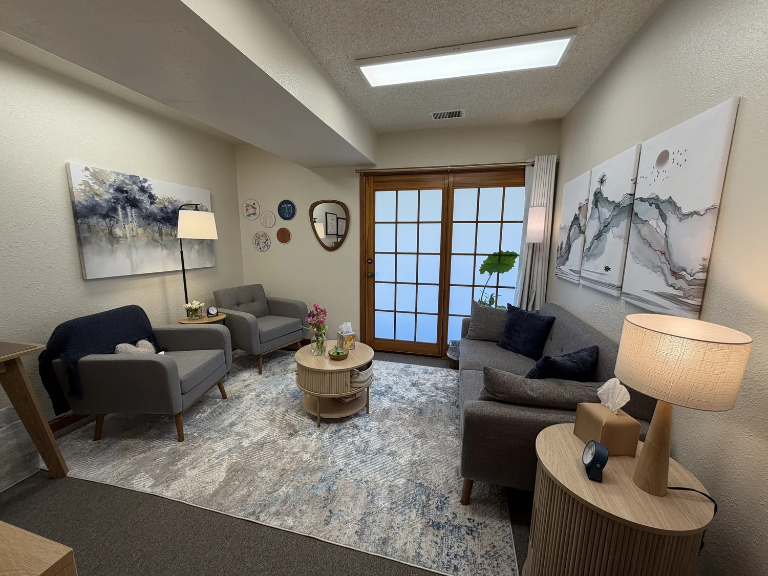 A cozy office with gray sofas, a wooden coffee table with flowers and tissues, a tall floor lamp, wall art and a potted plant.